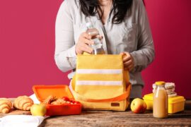 A close-up of someone's hands while packing snacks into a lunch box bag. They have apples, water, juice, and a sandwich.
