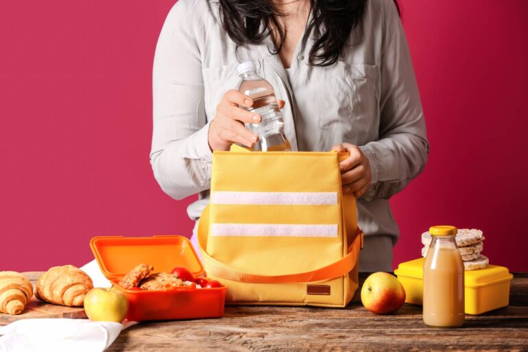A close-up of someone's hands while packing snacks into a lunch box bag. They have apples, water, juice, and a sandwich.