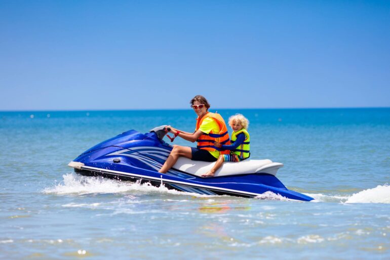 A father and a child ride on the back of a blue jet ski on a big body of water while wearing bright neon life vests.