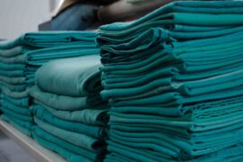 Four large stacks of folded green surgical fabrics and clothing on top of a large table inside an industrial laundry room.