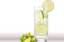 A tall glass of water with three slices of lime inside sits on a counter. Behind it, a white background is visible.