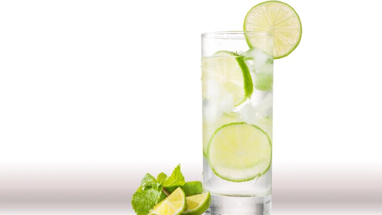 A tall glass of water with three slices of lime inside sits on a counter. Behind it, a white background is visible.