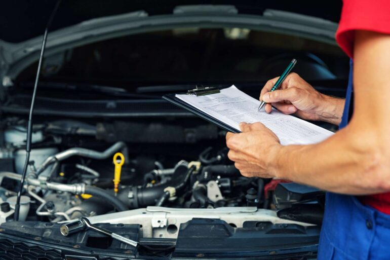 An automotive mechanic wearing a blue and red uniform holding a clipboard while inspecting a vehicle’s engine bay.