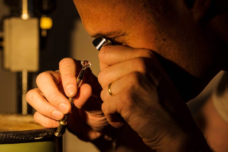 Close-up of a man closely examining a precious stone. He is holding it with a dop stick and using a magnifying loupe.