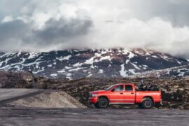 An orange pickup truck drives down a rural road with mountains, snow, and lots of clouds in the background.