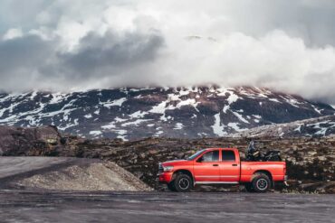An orange pickup truck drives down a rural road with mountains, snow, and lots of clouds in the background.