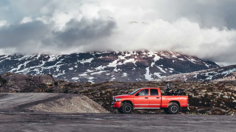 An orange pickup truck drives down a rural road with mountains, snow, and lots of clouds in the background.