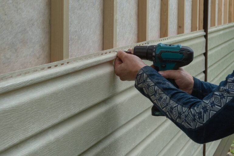 A person installs white siding on the side of their house while using a black and green cordless drill.