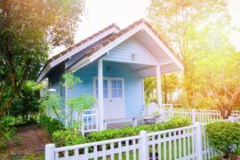 A blue tiny home sits in a green, forest area. There are white features, like the front door and a white fence.