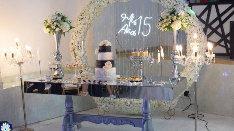 An elegant dessert table that has a cake, cupcakes, and other desserts on it as well as floral arrangements for a quince.