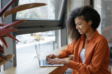 A young woman in an orange dress shirt smiling as she sits at a cafe table by a window and types on her laptop.