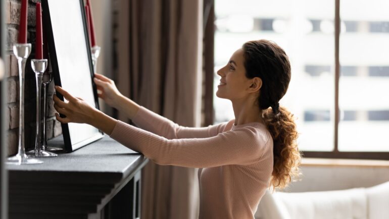 A woman decorating her living room. She is positioning a framed picture on her fireplace between decorative candles.