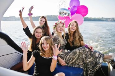 A group of young women having a bachelorette party on a boat. The bride to be is wearing a more colorful dress and a crown of flowers.