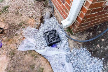 A plastic catch basin sits underneath a gutter spout on the corner of a home. Around the catch basin are lots of rocks.