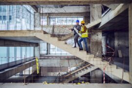 A construction worker in a work vest and an architect in a suit both wear hard hats and climb stairs on a construction site.