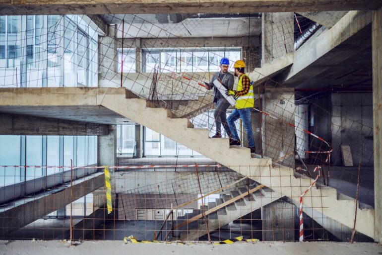 A construction worker in a work vest and an architect in a suit both wear hard hats and climb stairs on a construction site.