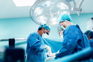 A team of medical professionals working at the surgery table under a big light, dressed in multiple layers of PPE.