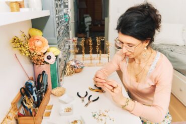 A woman sitting at a desk in her bedroom, creating handmade earrings using gold metal, multi-colored gems, and pliers.