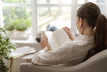 A woman sits in a chair at home and reads a book while relaxing. She has her feet on the windowsill.