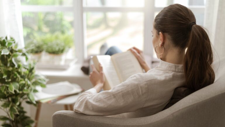A woman sits in a chair at home and reads a book while relaxing. She has her feet on the windowsill.