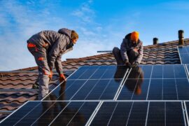 Two workers in orange and gray winter work gear working on top of a roof to install a set of solar panels.
