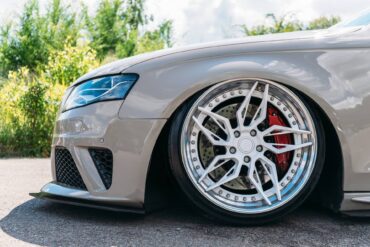 A close-up view shows a front driver's side tire on a beige vehicle parked on asphalt. Bushes appear behind the car.