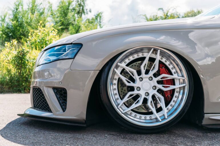 A close-up view shows a front driver's side tire on a beige vehicle parked on asphalt. Bushes appear behind the car.