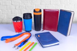 A collection of promotional products including thermos mugs, lanyard neck straps, pens, and notebooks are on a desk.