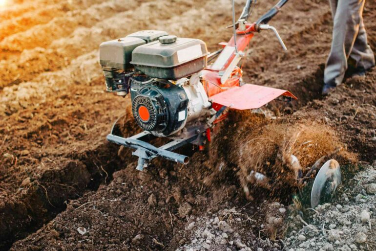 Someone who homesteads running a hand tiller through their land. They're wearing long pants and work boots.