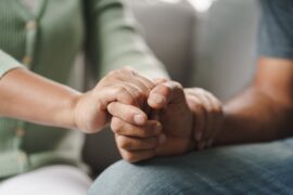 A close up shot of two people holding hands in an office setting. One person's hand is also on the other's arm.