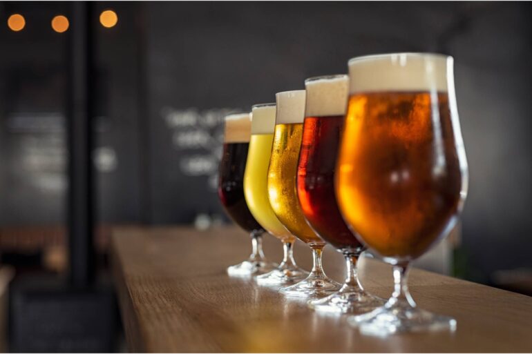 Close-up of five tall glasses filled with different types of beers with varied colors sitting on top of a wooden counter.