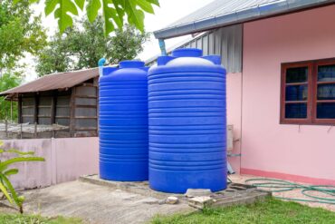 Two tall blue plastic water tanks on a concrete platform beside a pink building, grass, and trees in the background.