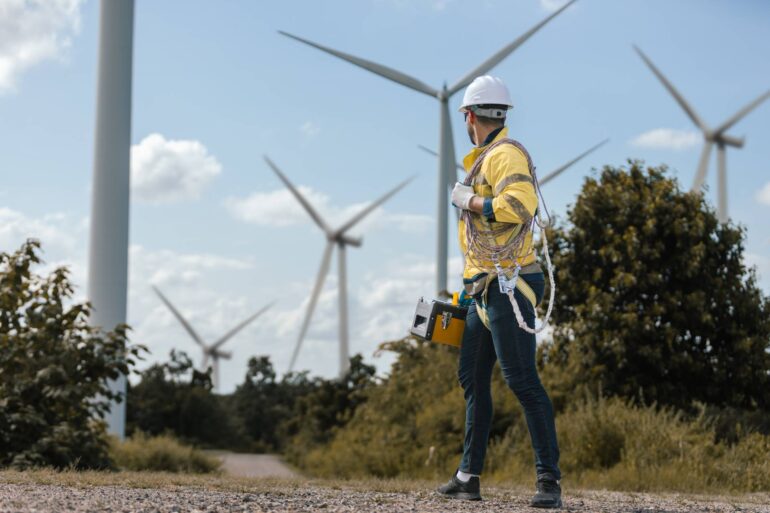 A wind turbine maintenance worker standing and looking back at wind turbines. He's wearing a helmet.