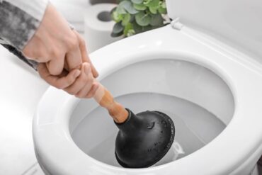 A close-up of a person's hands using a plunger in a toilet. There is a roll of toilet paper and a plant near the toilet.