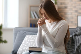 A woman in a white sweater sits on a gray couch, bowing her head and folding her hands. A book sits in her lap.