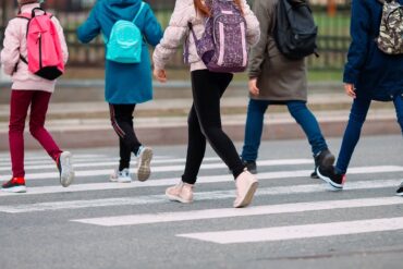 A group of young students walking across the street on the crosswalk. They're in coats with backpacks.