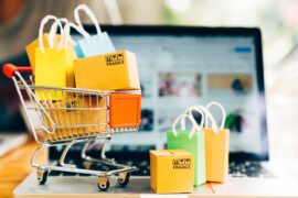A mini shopping cart sitting on a laptop's keyboard with small, yellow shopping bags in and out of the cart.