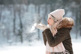 A woman in a brown coat and pink hat and scarf blows snow out of her hands while standing outside.