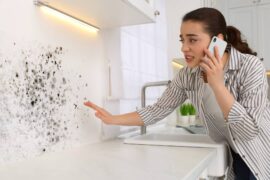 A woman looking at mold on a wall in the kitchen. She is on her phone and has a concerned look on her face.