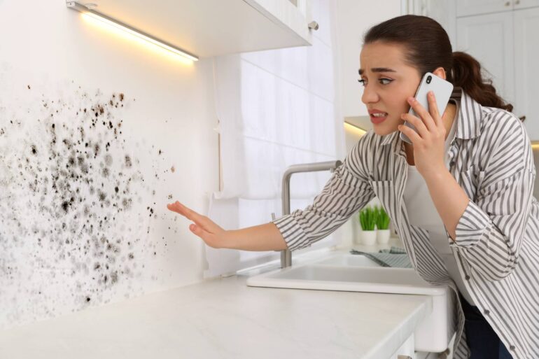 A woman looking at mold on a wall in the kitchen. She is on her phone and has a concerned look on her face.