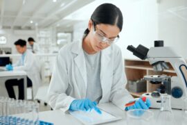 A woman in a lab coat is looking at something on a device. There are two people in the background also wearing lab coats.