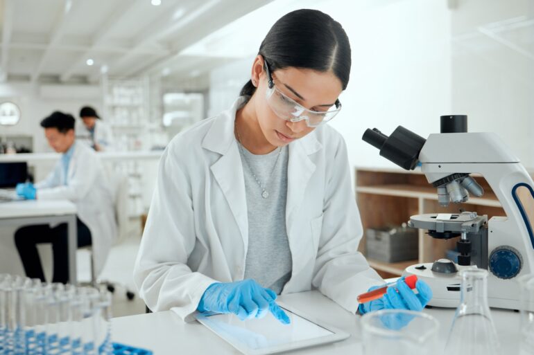 A woman in a lab coat is looking at something on a device. There are two people in the background also wearing lab coats.