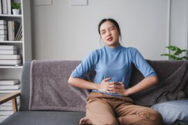 A woman sitting on her couch holds her stomach and winces in pain. She wears a blue shirt and tan pants.