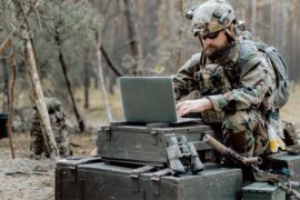 A soldier using a laptop on a makeshift desk of boxes in the woods. He is wearing a backpack and shades.