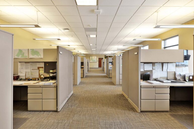 The walkway of an office with cubicles on both sides. They have papers on the desks and images pinned on the walls.
