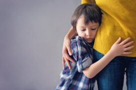 A young boy holds on to his mother's side and lays his head on her hip as she holds him close with a hand on his back.