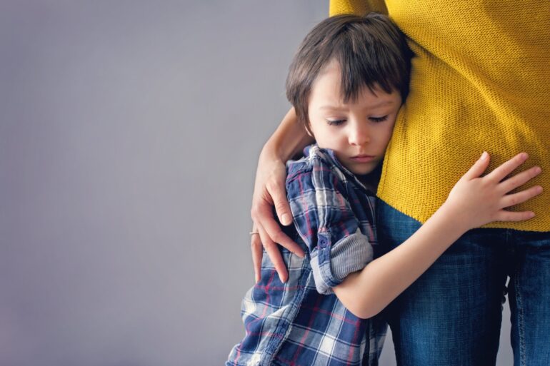A young boy holds on to his mother's side and lays his head on her hip as she holds him close with a hand on his back.