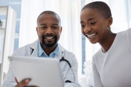 A doctor in a holds a tablet in front of a patient. The doctor and the patient are both smiling.