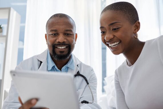 A doctor in a holds a tablet in front of a patient. The doctor and the patient are both smiling.