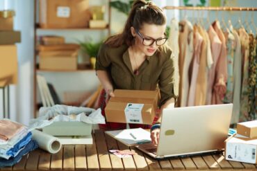 A woman in an olive shirt and glasses looking at a laptop while holding a box with a shipping label on it.